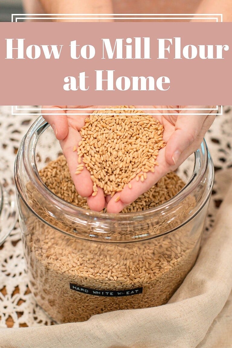 How to Mill Flour at Home text overlay above cupped hands holding hard white wheat berries over a glass jar filled with grain, set on a lace-topped table with a linen cloth