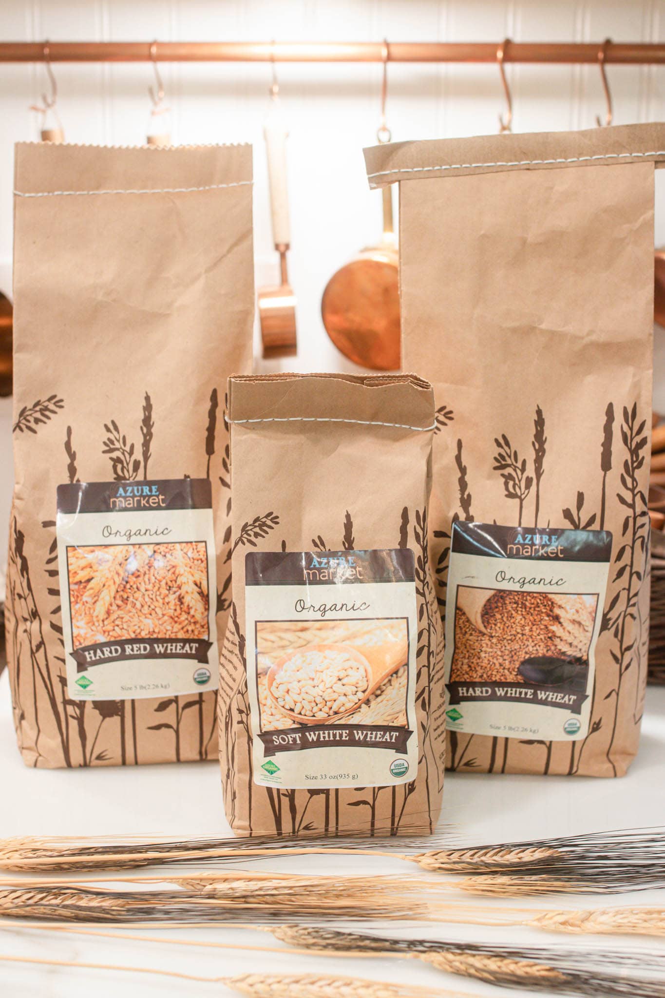 Bags of organic wheat berries labeled hard red wheat, soft white wheat, and hard white wheat standing on a kitchen counter with wheat stalks placed in front.