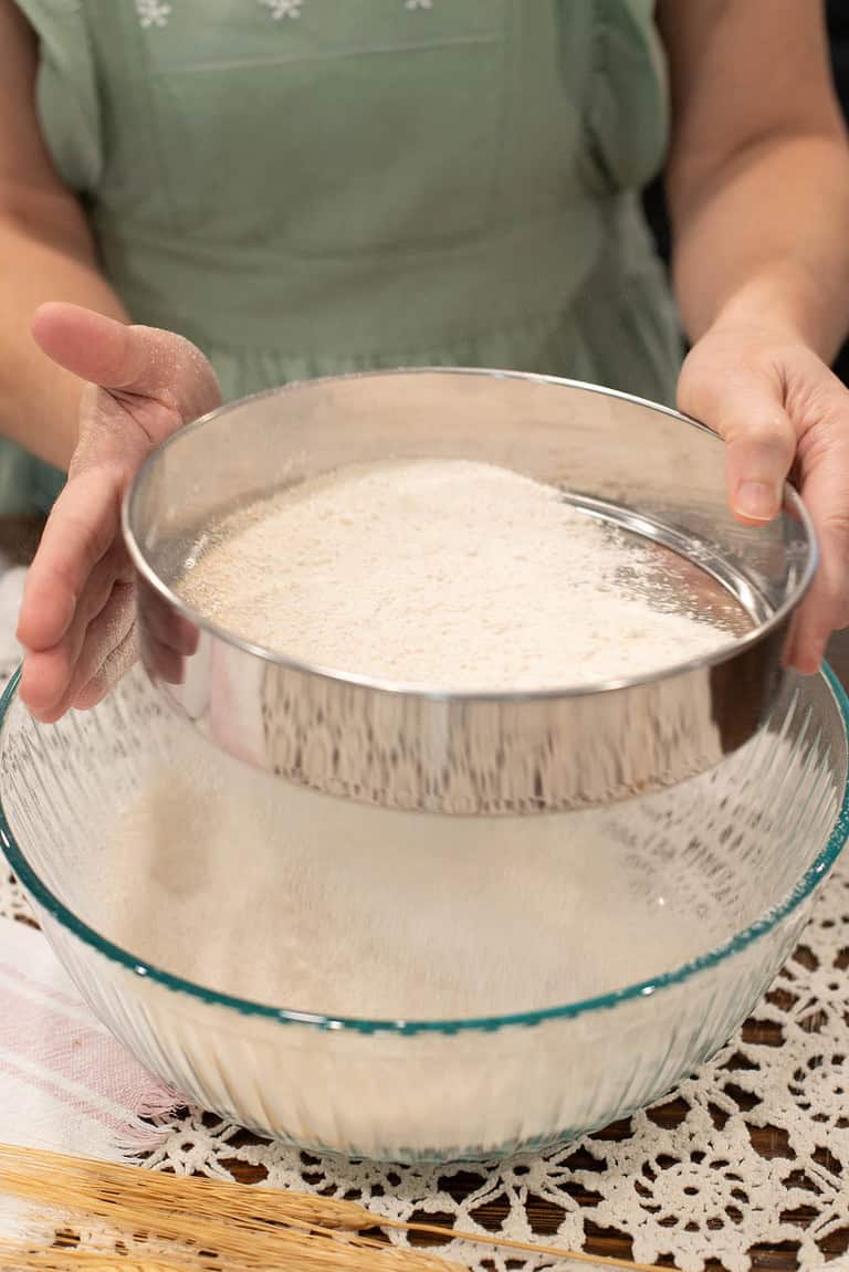 Person in a green dress manually sifting freshly milled flour over a glass bowl using a round stainless steel sieve.