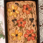 Golden baked sourdough focaccia made with freshly milled flour, decorated with cherry tomatoes, olives, and rosemary in a cast iron pan.