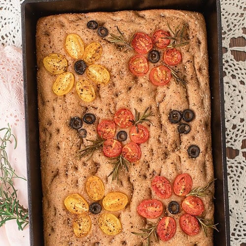 Golden baked sourdough focaccia made with freshly milled flour, decorated with cherry tomatoes, olives, and rosemary in a cast iron pan.