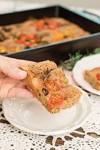 Hand holding a slice of sourdough focaccia made with freshly milled flour, topped with cherry tomatoes, olives, and rosemary, with a baking pan in the background.