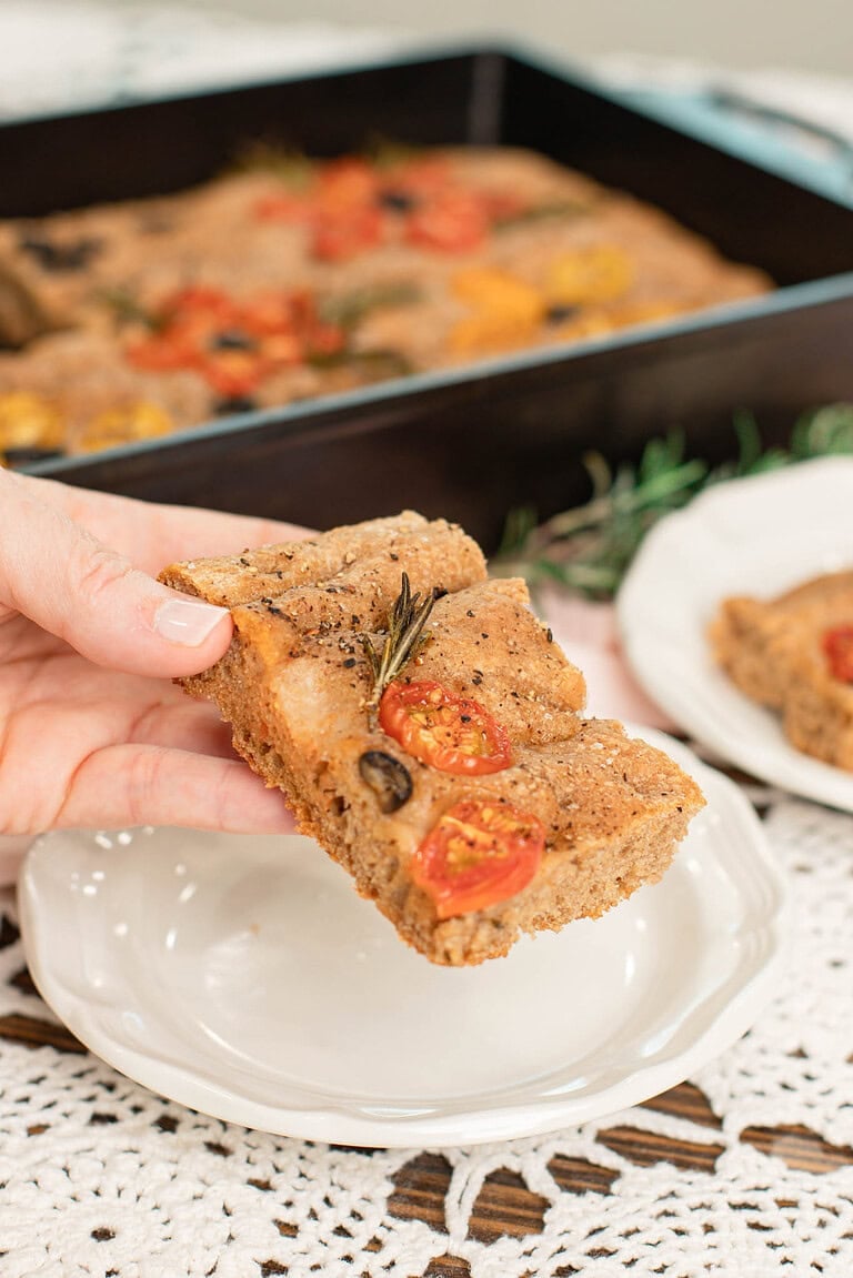 Hand holding a slice of sourdough focaccia made with freshly milled flour, topped with cherry tomatoes, olives, and rosemary, with a baking pan in the background.