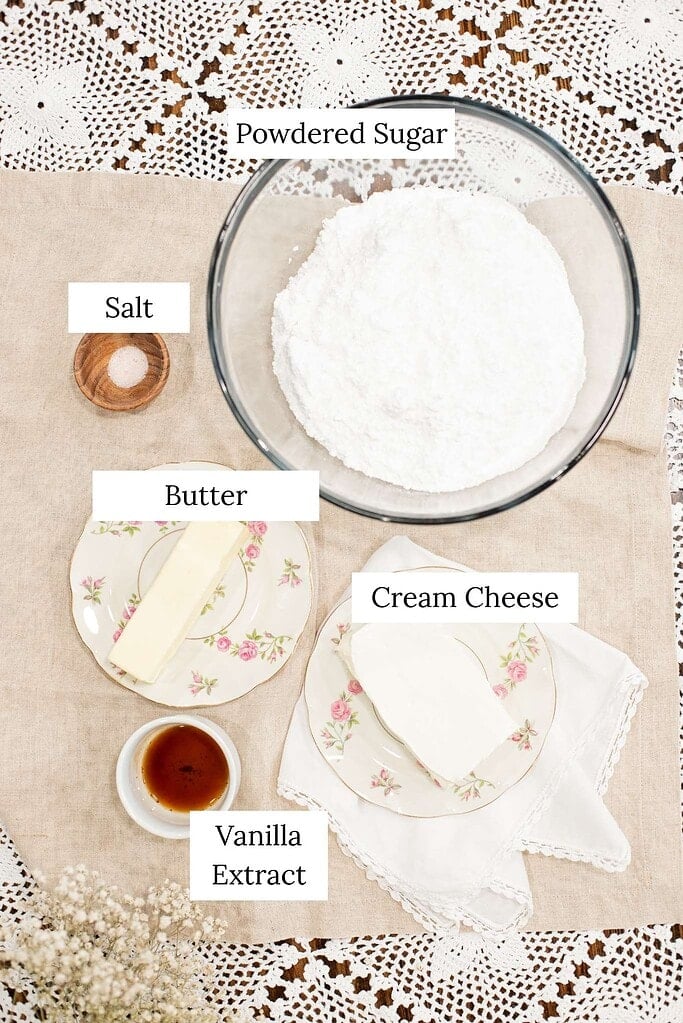 Overhead view of cream cheese frosting ingredients including powdered sugar, butter, cream cheese, vanilla extract, and salt arranged on a linen cloth.