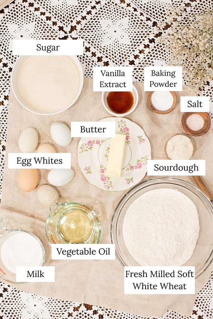 Overhead view of sourdough vanilla cake ingredients including fresh milled soft white wheat flour, sugar, egg whites, milk, vegetable oil, butter, sourdough starter, baking powder, salt, and vanilla extract.