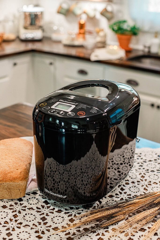 Black bread machine on a kitchen counter next to a loaf of bread and wheat stalks, used for baking bread with freshly milled flour.