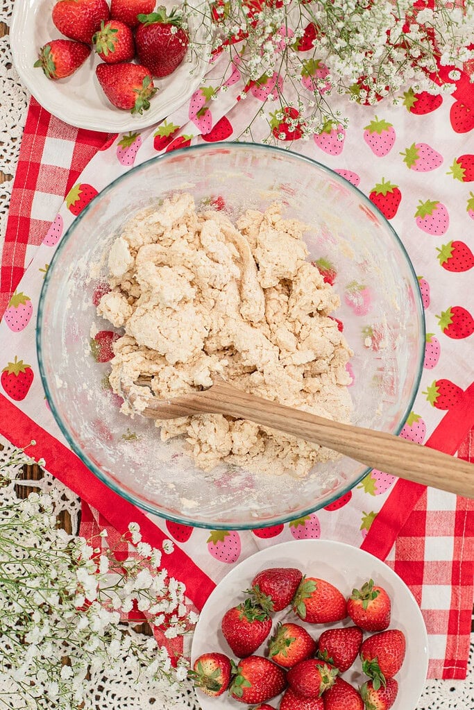Freshly milled flour dough being mixed in a glass bowl with a wooden spoon, surrounded by fresh strawberries and a strawberry-patterned cloth.