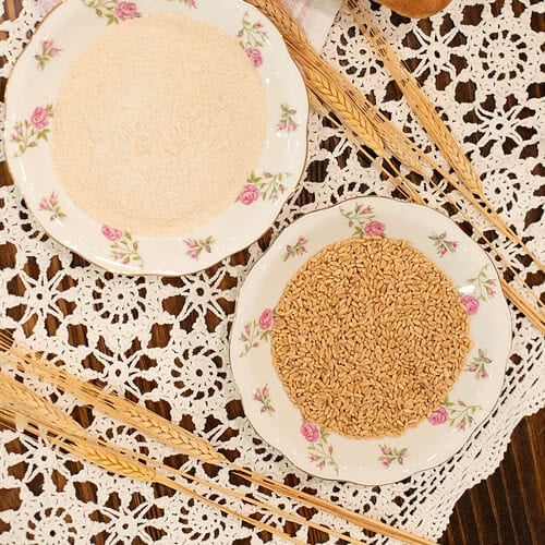 Overhead view of a loaf of freshly baked whole wheat bread on a pink-striped cloth napkin. Below it are two floral china plates—one filled with freshly milled whole wheat flour, and the other with whole wheat berries—set atop a vintage lace tablecloth with decorative wheat stalks arranged nearby.