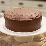 A slightly angled close-up of the fully frosted cake, showing the smooth, even chocolate buttercream and clean cake edges. Baby’s breath and lace set the scene.