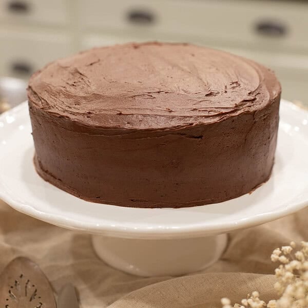 A slightly angled close-up of the fully frosted cake, showing the smooth, even chocolate buttercream and clean cake edges. Baby’s breath and lace set the scene.