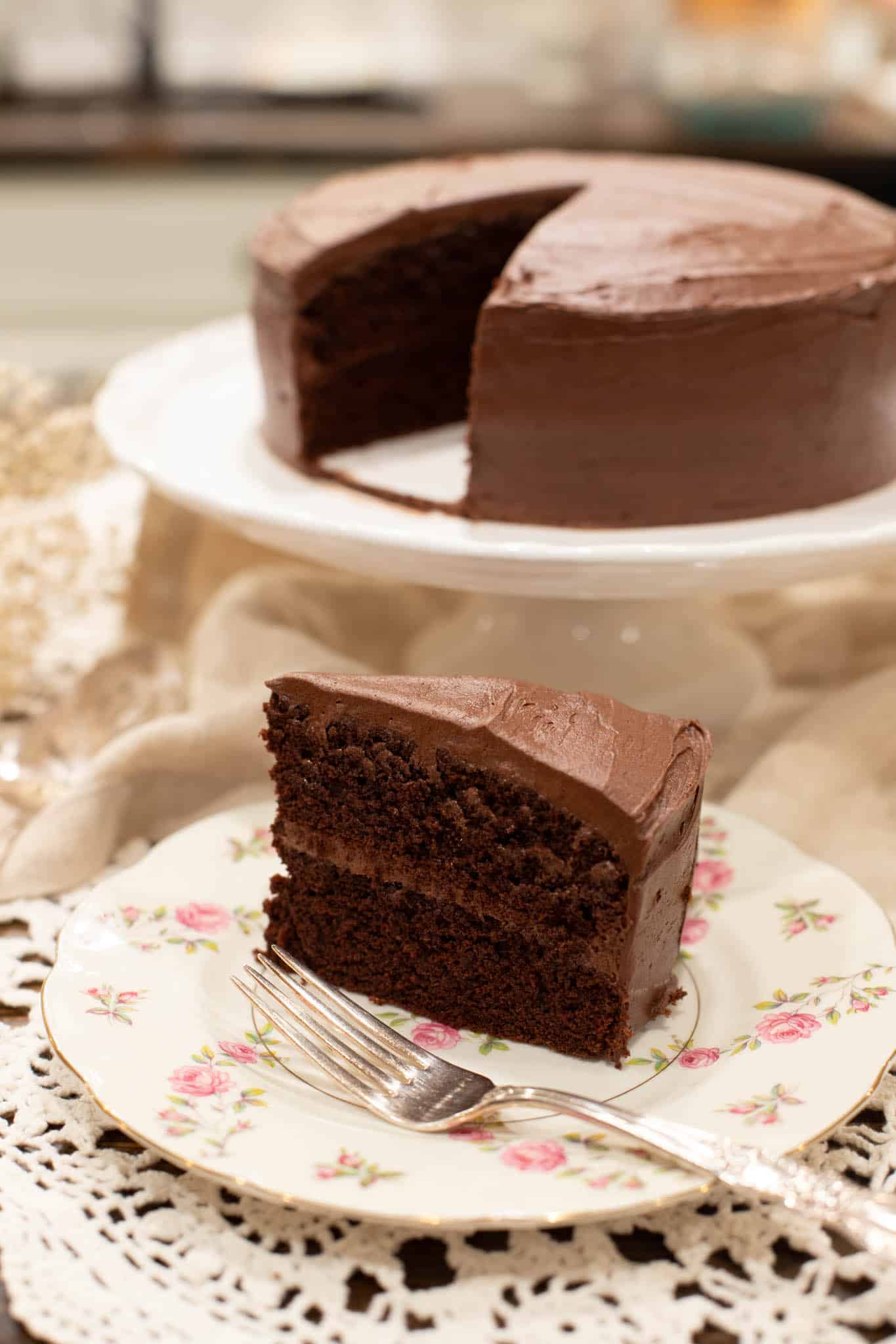 The plated slice of chocolate cake sits ready to enjoy on a vintage floral plate with a silver fork. The full cake in the background shows where the slice was removed.