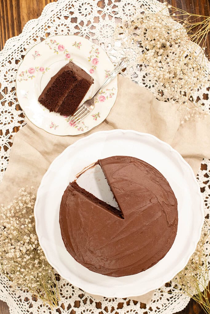 An overhead shot of the chocolate cake with one slice removed and plated nearby. The cake is on a white pedestal surrounded by dried flowers and crochet lace for a nostalgic, cozy touch.
