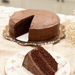 A close-up of a slice of rich chocolate sourdough cake with thick, velvety frosting, served on a vintage floral plate. The full frosted cake with a slice missing is visible in the background on an ornate silver platter.