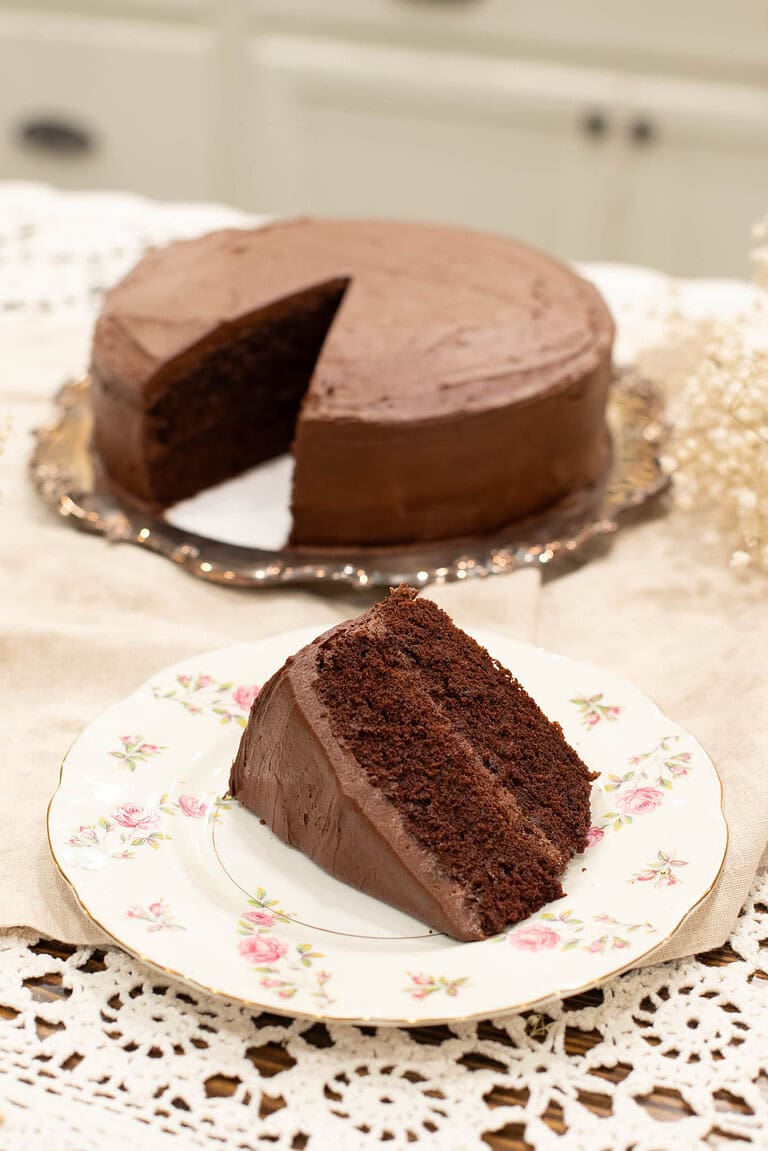 A close-up of a slice of rich chocolate sourdough cake with thick, velvety frosting, served on a vintage floral plate. The full frosted cake with a slice missing is visible in the background on an ornate silver platter.