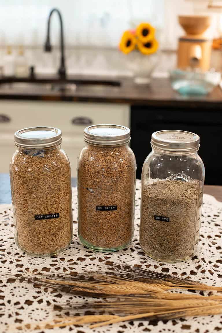 Three labeled mason jars filled with various grains—Oat Groats, OG Spelt, and OG Rye—sit on a lace doily on a kitchen counter, with a bundle of wheat stalks in front and a kitchen sink in the background.