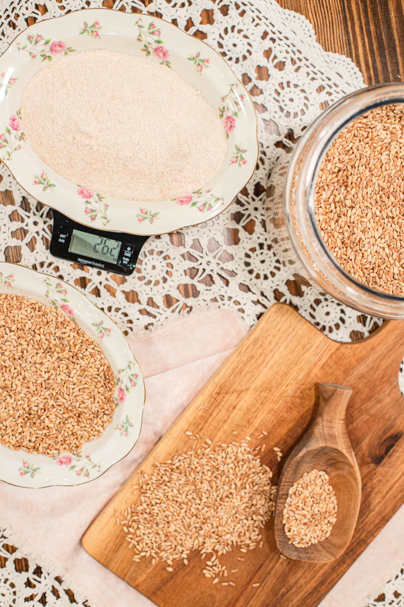 Freshly milled flour in a floral bowl on a digital scale surrounded by whole wheat berries, a wooden scoop, and a cutting board on a lace tablecloth