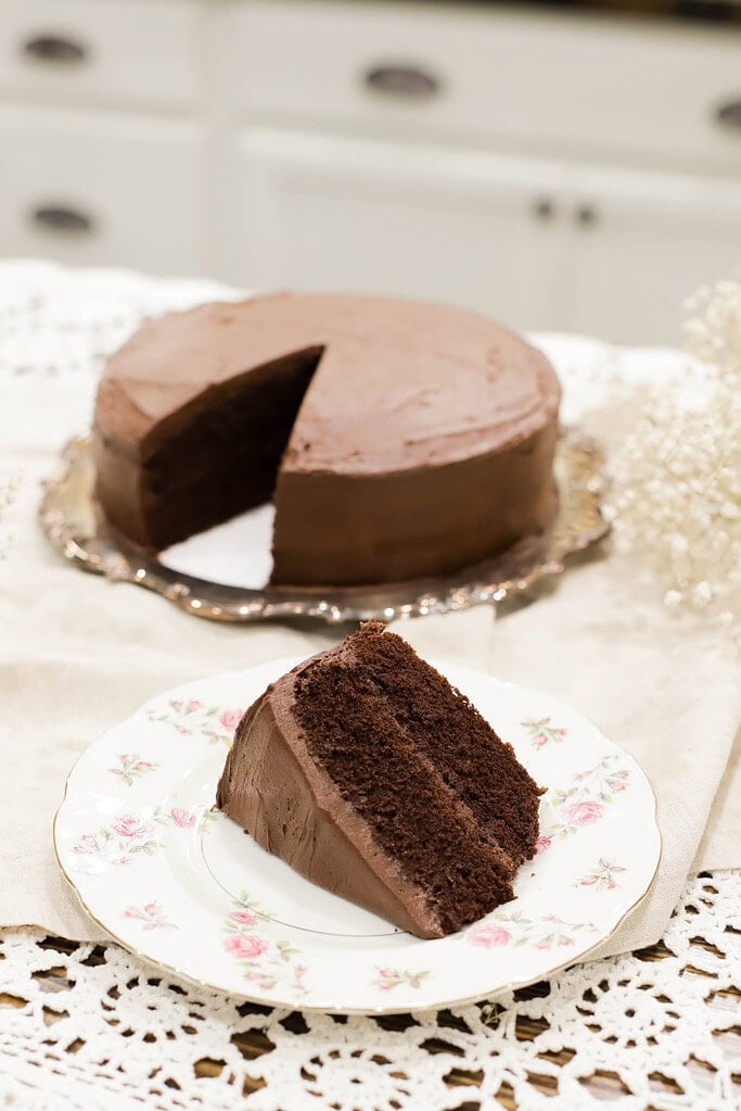 Slice of chocolate layer cake with smooth chocolate frosting on a floral plate, with the full cake in the background on a serving tray
