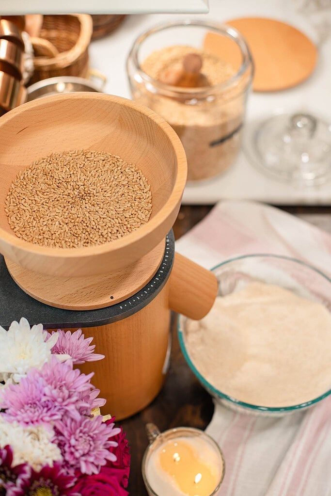 Whole wheat berries in a wooden bowl on top of a countertop grain mill with freshly milled flour nearby