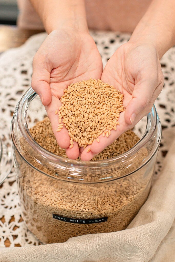 Hands holding wheat berries above a glass jar labeled hard white wheat on a lace tablecloth