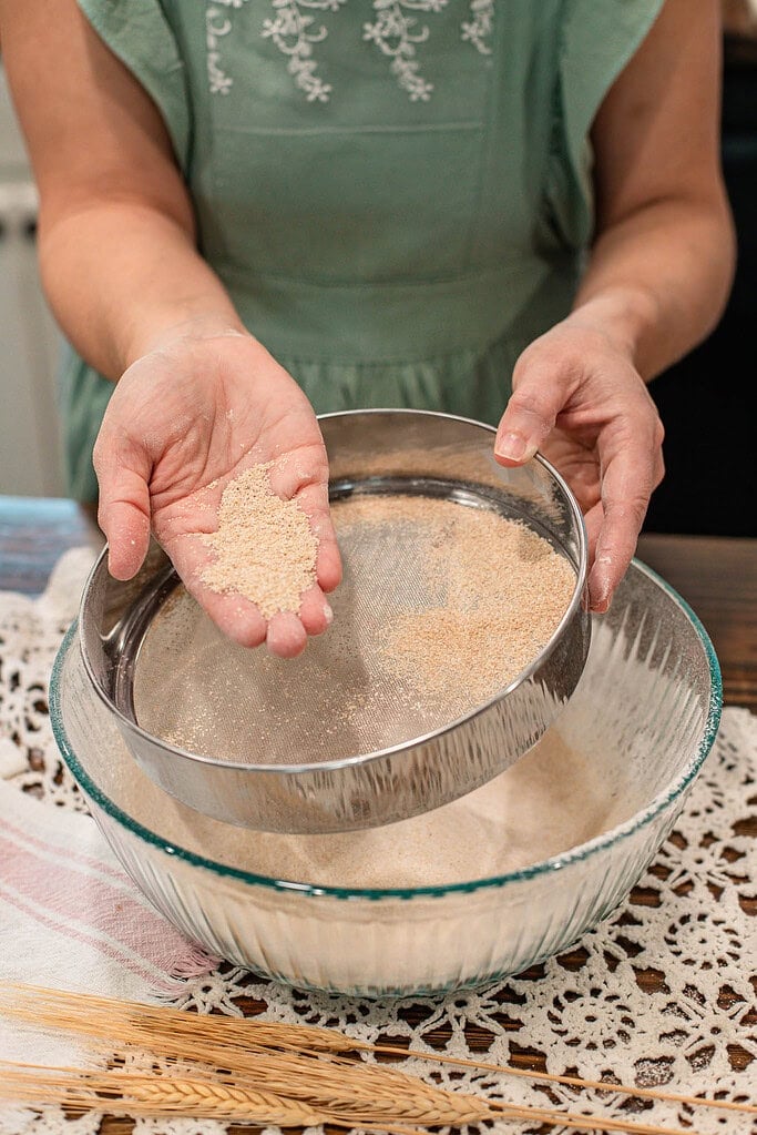 Hand holding coarse bran separated from freshly milled flour in a sieve above a glass bowl, showing texture difference after sifting.