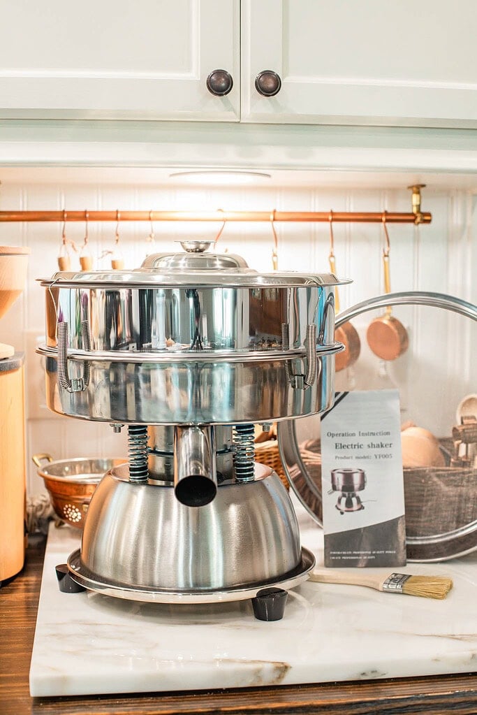 Electric flour sifter machine on a marble countertop in a cozy cottage kitchen with copper utensils hanging in the background.