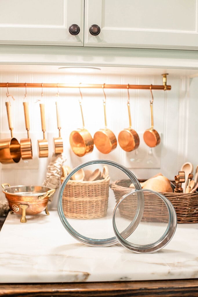 Two fine mesh flour sieves resting on a marble countertop with copper measuring cups and woven baskets in a cottage-style kitchen.