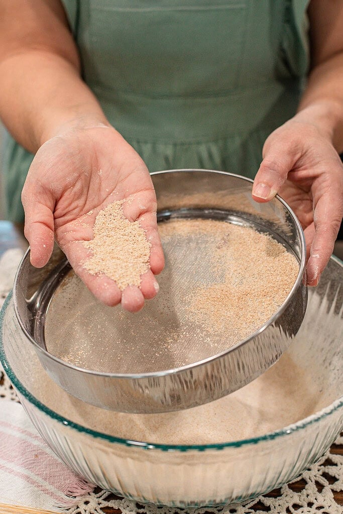 Hand holding coarse bran separated from freshly milled flour in a sieve above a glass bowl, showing texture difference after sifting.