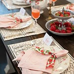 Angled view of a place setting featuring a pink napkin tied with strawberry-printed ribbon on a floral plate atop a lace placemat.