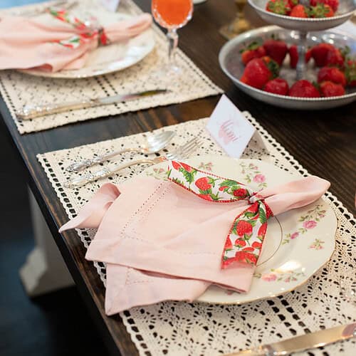 Angled view of a place setting featuring a pink napkin tied with strawberry-printed ribbon on a floral plate atop a lace placemat.