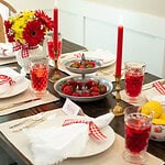 Warm summer tablescape featuring a bright bouquet of red and yellow flowers, red gingham napkins, a tray of fresh strawberries, and vintage glassware under cozy candlelight.