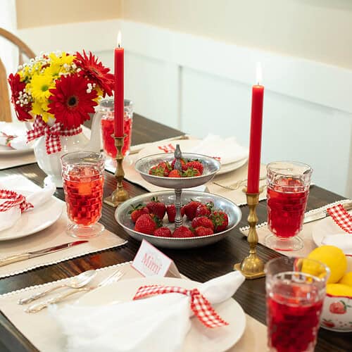 Warm summer tablescape featuring a bright bouquet of red and yellow flowers, red gingham napkins, a tray of fresh strawberries, and vintage glassware under cozy candlelight.