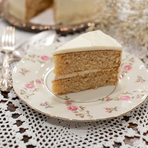 A beautifully plated slice of vanilla cake with buttercream frosting, with a blurred background of the remaining cake and delicate baby’s breath flowers.