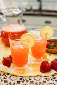 Two glasses of strawberry blush lemonade garnished with lemon slices and filled with ice, sitting on a yellow gingham cloth with fresh strawberries beside them and a glass pitcher in the background.