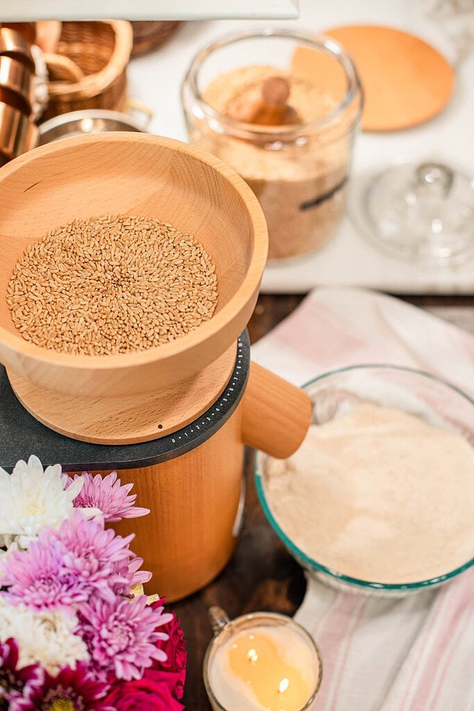 Wooden bowl filled with whole wheat berries sitting on top of a countertop grain mill, with freshly milled flour in a glass bowl nearby, surrounded by soft flowers and a lit candle in a cozy kitchen setting.