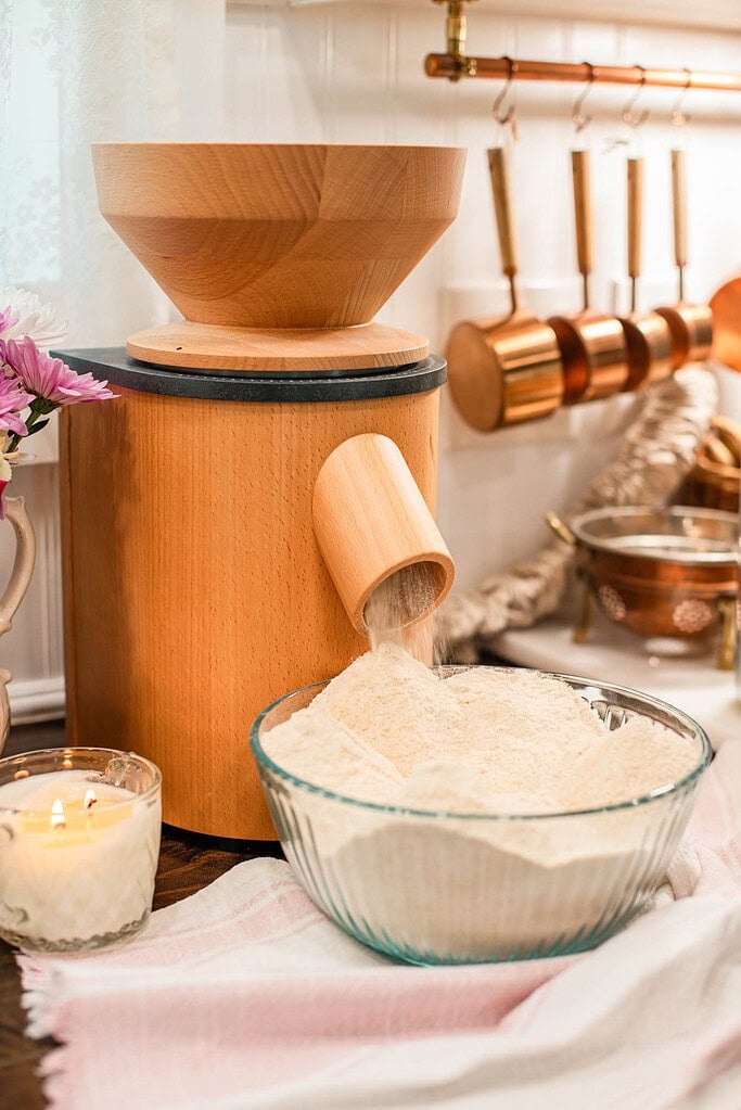 Freshly milled flour pouring out of a wooden grain mill into a clear glass mixing bowl, with copper kitchen tools hanging in the background and a candle glowing beside it.