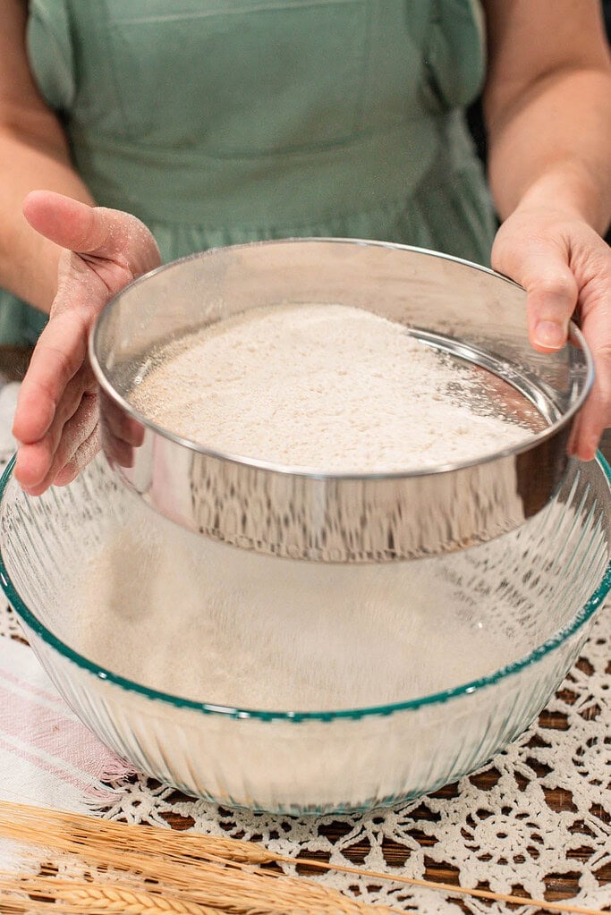 Hands holding a fine mesh sieve over a large glass bowl, sifting freshly milled flour to separate bran for a lighter texture, with a lace tablecloth underneath.