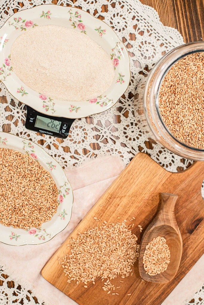 Overhead view of wheat berries and freshly milled flour measured on a kitchen scale, arranged on a wooden board and lace cloth with a glass jar of grains nearby.