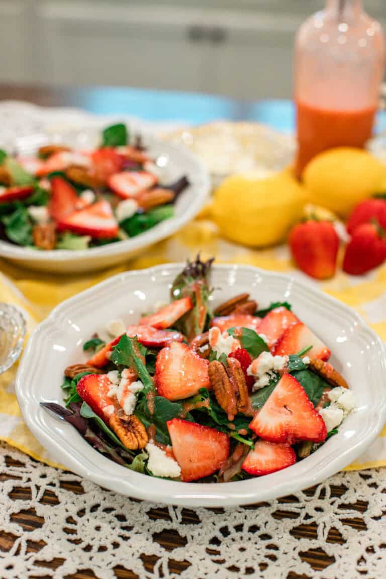 Slightly angled shot of two strawberry salads with pecans and feta on a yellow gingham cloth. A vintage glass dressing bottle and fresh strawberries and lemons complete the cozy summer table setting.