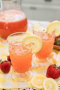 Close-up of two glasses of pink strawberry lemonade over ice, garnished with lemon slices, with fresh strawberries on the table.