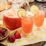Pitcher of strawberry lemonade with two filled glasses garnished with lemon slices and fresh strawberries on a yellow gingham cloth.