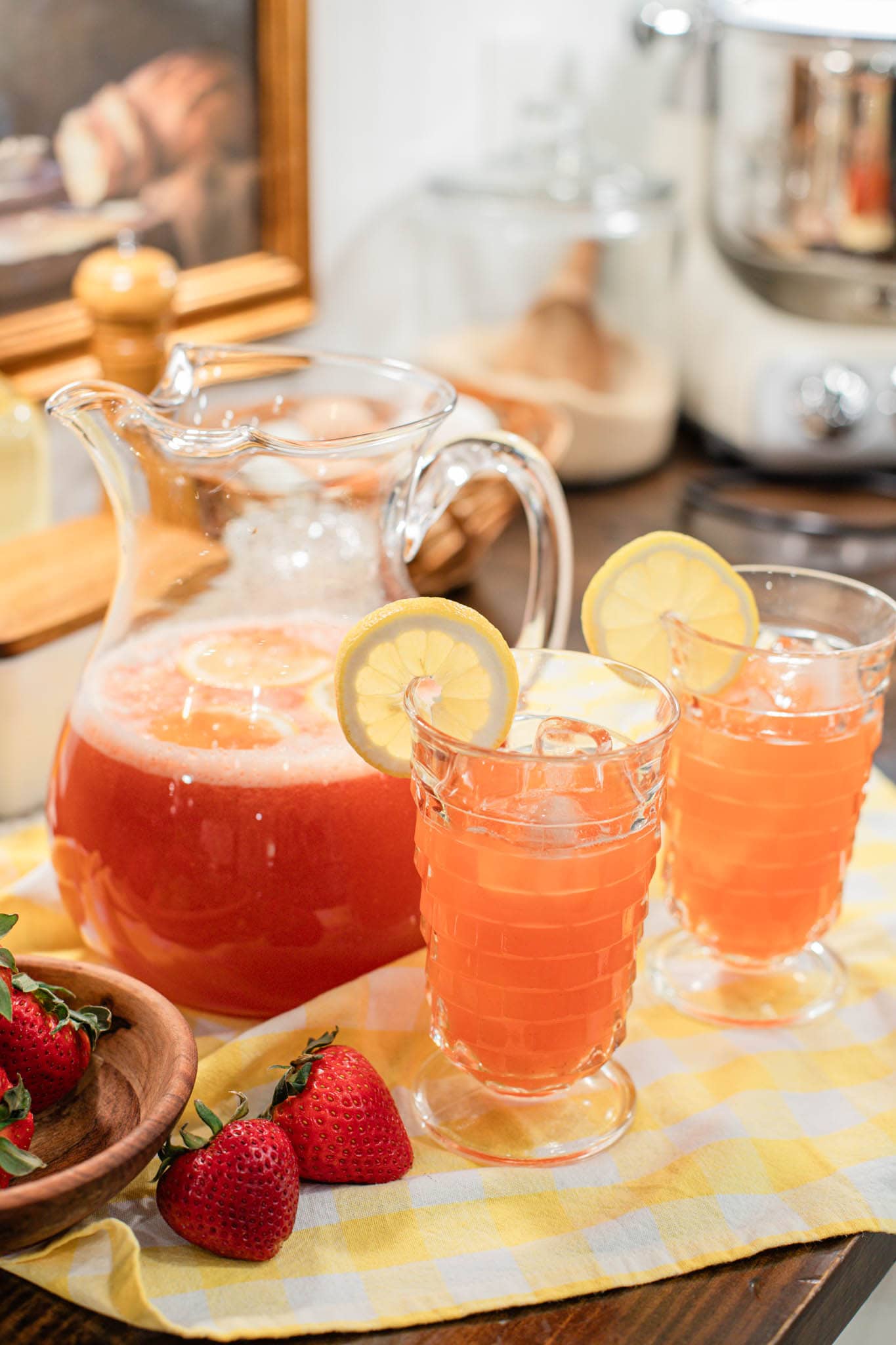 Pitcher of strawberry lemonade with two filled glasses garnished with lemon slices and fresh strawberries on a yellow gingham cloth.