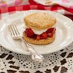 Assembled biscuit-style strawberry shortcake with whipped cream and juicy macerated strawberries on a white vintage plate with a silver fork.