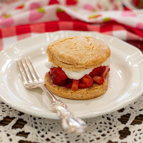 Assembled biscuit-style strawberry shortcake with whipped cream and juicy macerated strawberries on a white vintage plate with a silver fork.