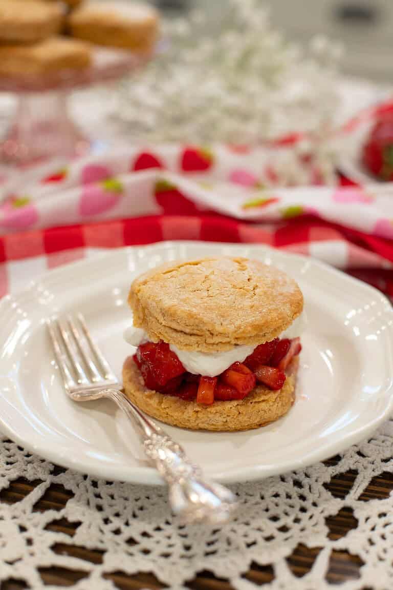 Assembled biscuit-style strawberry shortcake with whipped cream and juicy macerated strawberries on a white vintage plate with a silver fork.