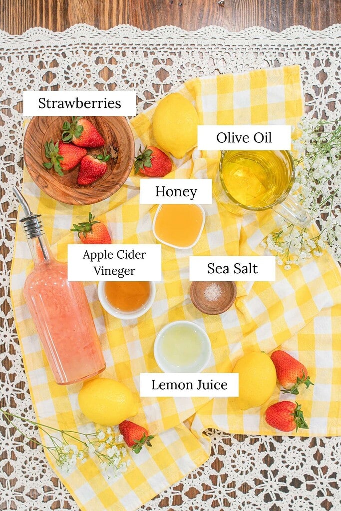 Ingredients for strawberry vinaigrette including fresh strawberries, olive oil, honey, apple cider vinegar, lemon juice, and sea salt arranged on a yellow gingham cloth.