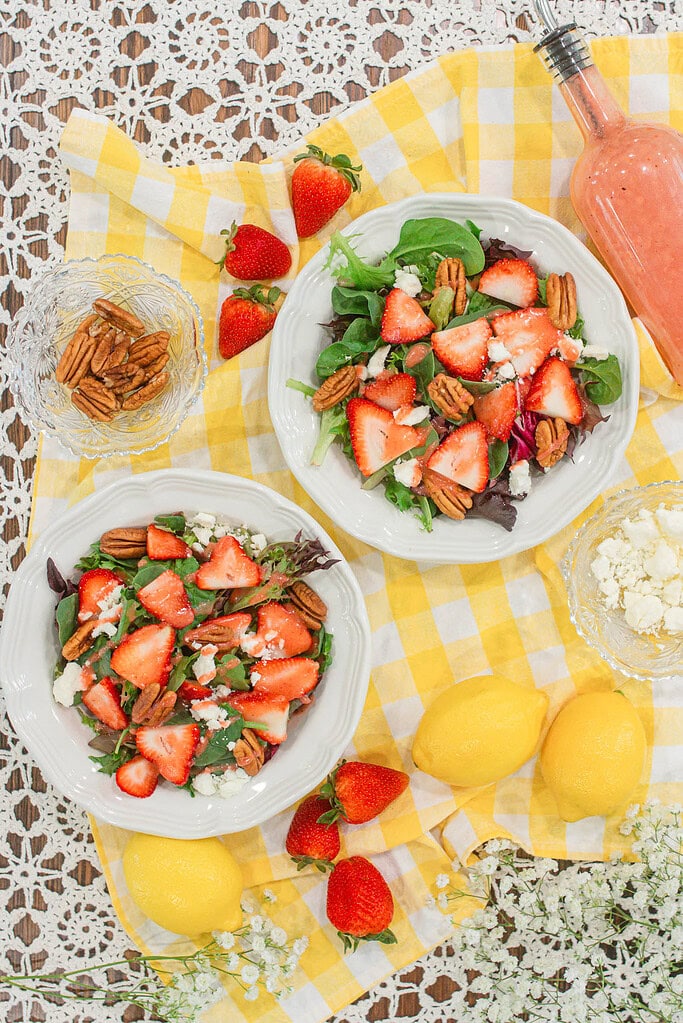 Overhead view of strawberry spinach salad bowls with fresh strawberries, pecans, feta cheese, and strawberry vinaigrette on a yellow gingham cloth.