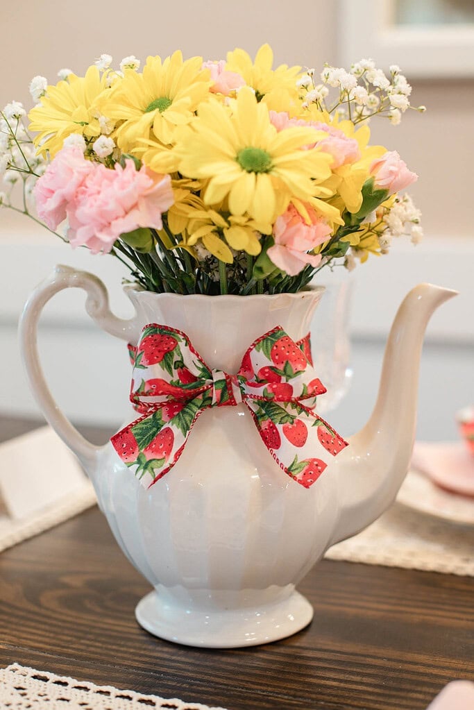 Floral arrangement in teapot with strawberry ribbon and yellow daisies on cottage table