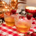 Two clear glass mugs of freshly steeped herbal tea garnished with strawberries, sitting on a red gingham cloth with a warm, candlelit copper kettle tea station in the background.