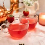 Two clear glass mugs filled with freshly brewed strawberry hibiscus tea, garnished with strawberries and nestled among baby’s breath and glowing candles on a pale pink cloth.