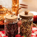Glass jars of loose leaf strawberry tea ingredients on red gingham cloth with fresh strawberries and copper teapot in background
